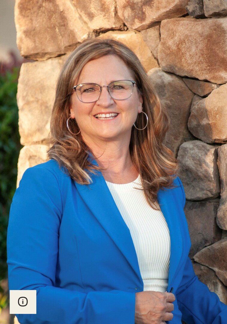 A woman in glasses and blue jacket standing next to a rock wall.